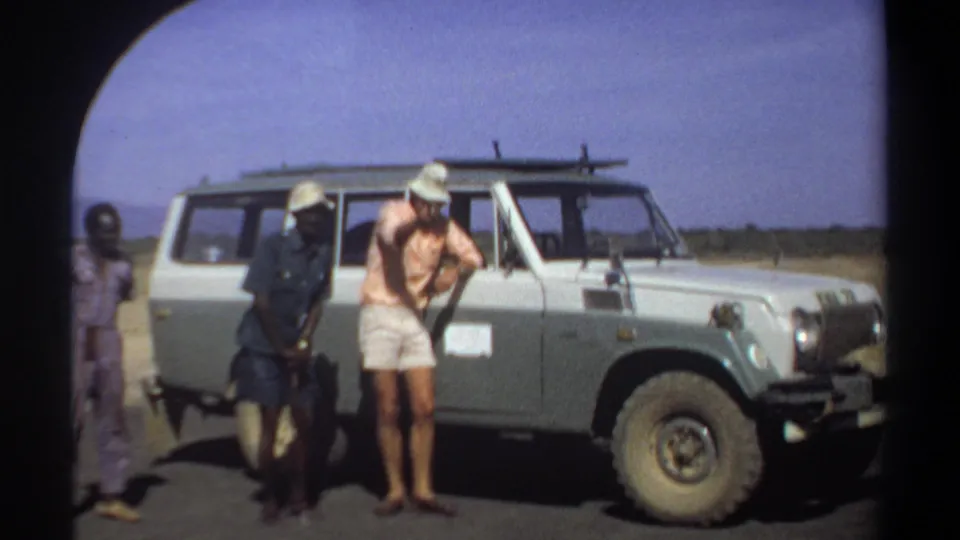 Beside the pale utility vehicle in 1969 Khartoum, a man in a pink shirt gestures while others stand nearby.