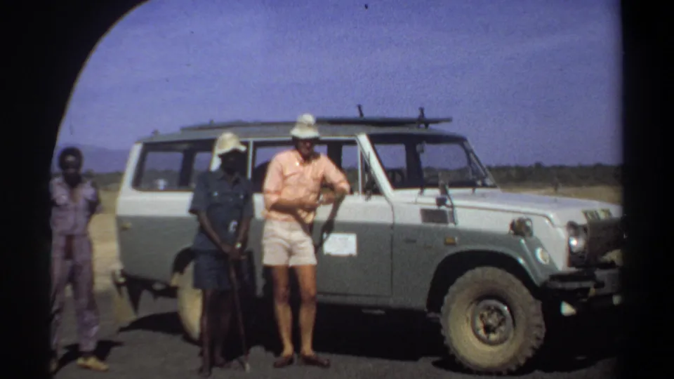 In the 1969 Khartoum footage, a man in shorts and a light hat stands with two local men beside the vehicle, one holding a walking stick.