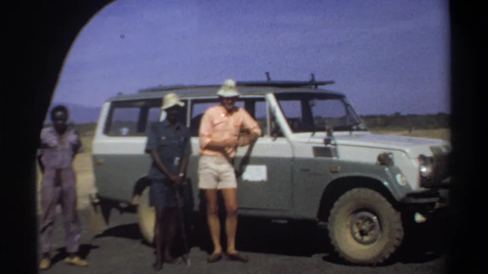 The 1969 Sudan home-movie frame shows the group standing close to the Land Cruiser, with the central man partly shaded and the visitor near the door.