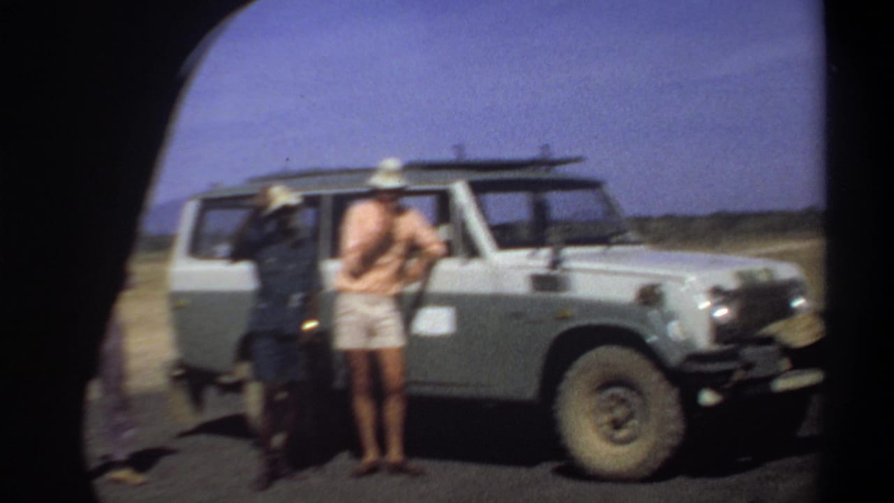Three adult men stand beside a white off-road vehicle on a paved road in an arid landscape.