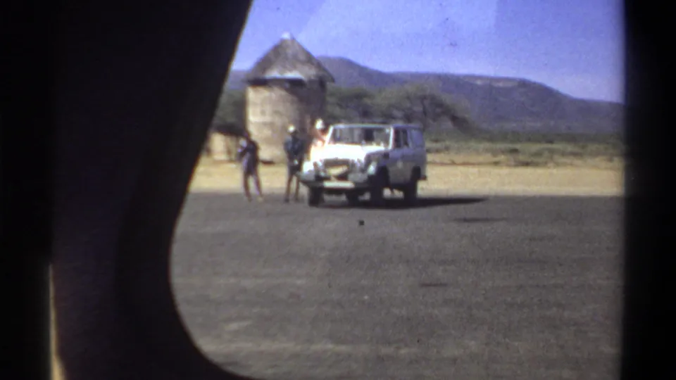 1969 Khartoum home-movie frame showing a white utility vehicle, several men, dry ground, trees, and round thatched structures.