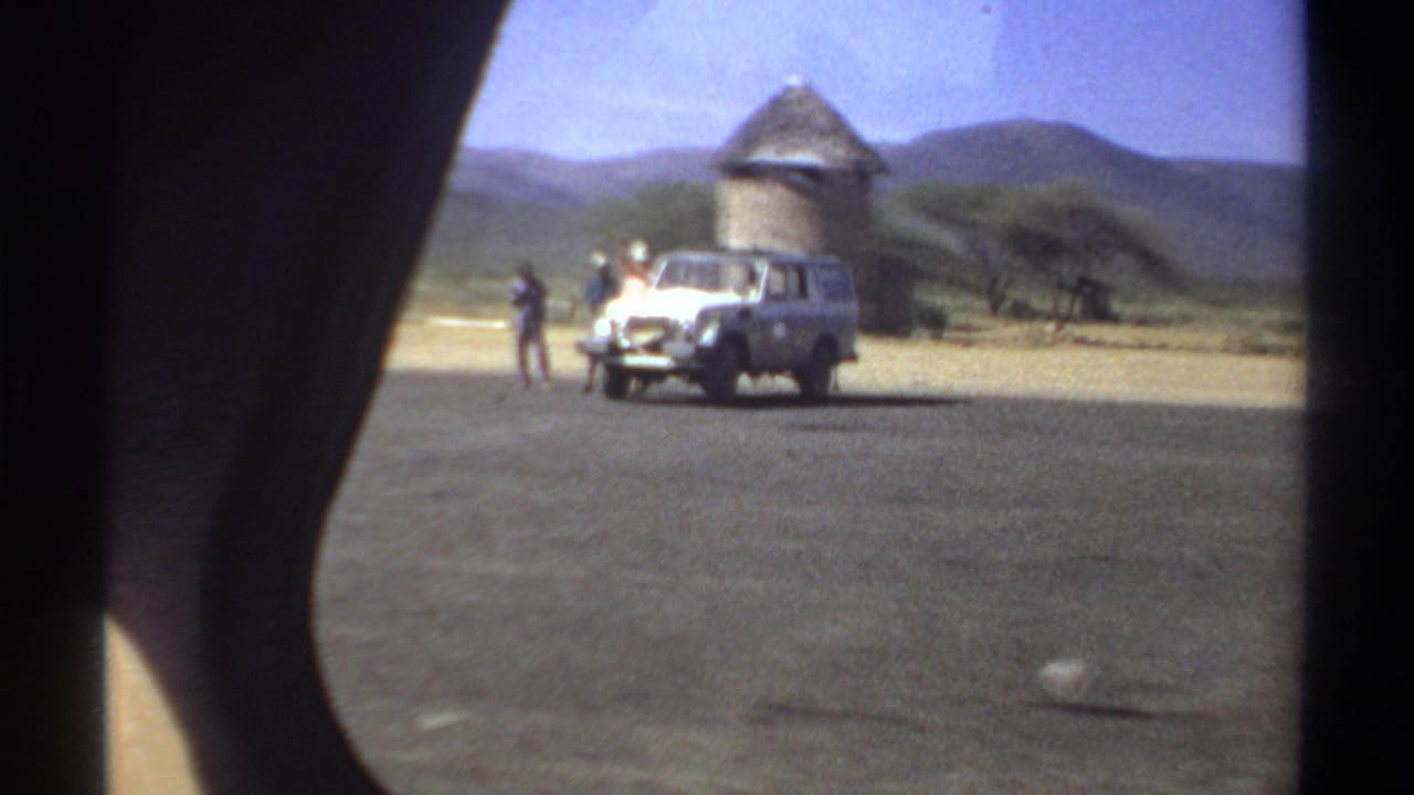 1969 Khartoum frame with a white utility vehicle, men standing nearby, a thatched round structure, trees, and low hills.