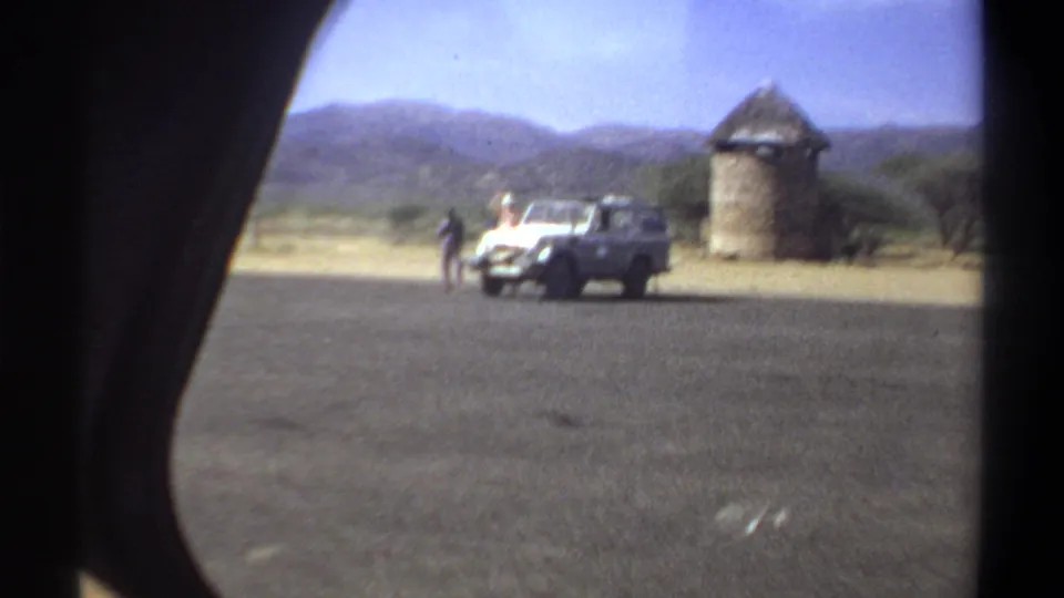 Moving-vehicle view in 1969 Khartoum showing a white utility vehicle, men, roadside pavement, and a round thatched structure.