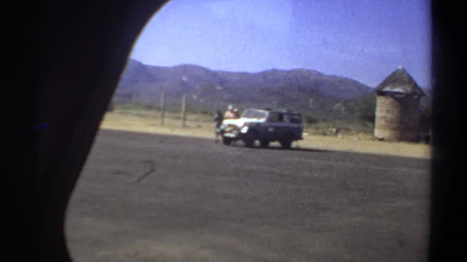 1969 Khartoum frame showing the white utility vehicle farther right, men near the roadside, hills, dry terrain, and a thatched structure.