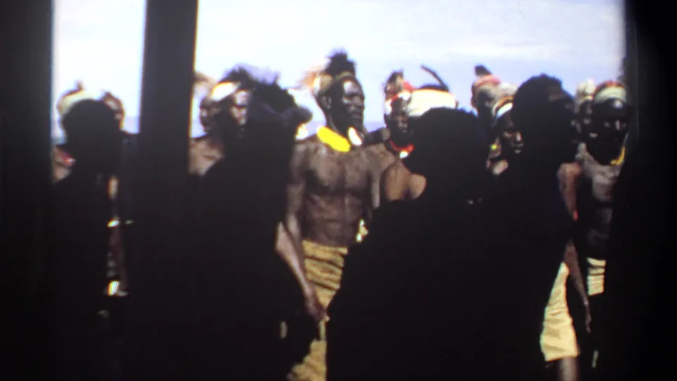 1969 Sudan home-movie frame showing a crowded outdoor group, with silhouetted figures close to the camera and dressed men behind.