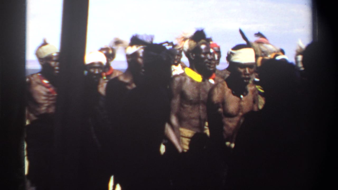 Men in traditional costume stand or move near water, partly blocked by dark foreground figures.