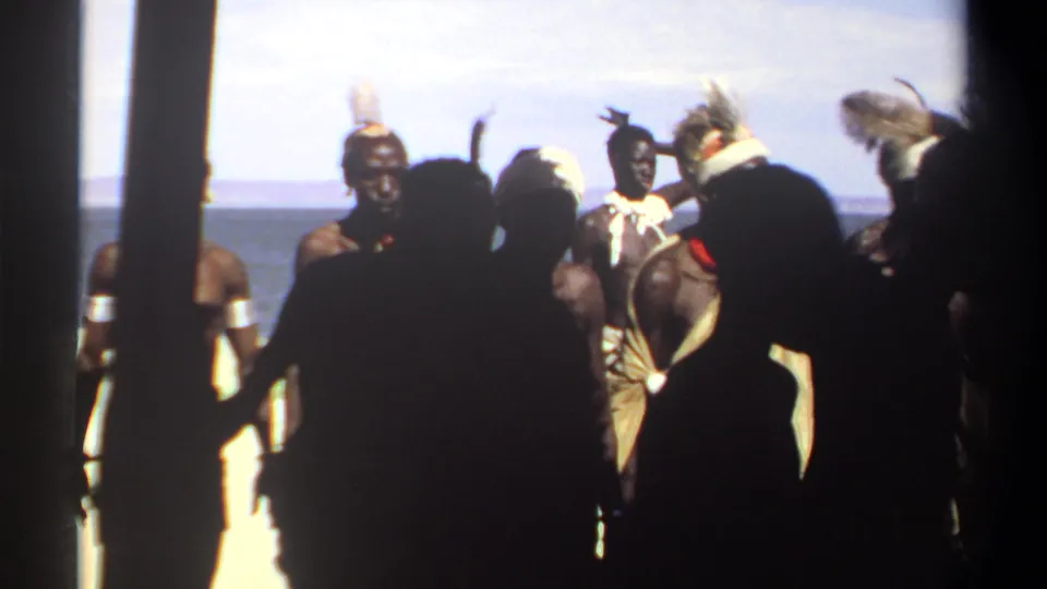 1969 Sudan 8mm frame where dark foreground figures partly block men wearing headbands, armbands, cloth wraps, and headdresses by the water.