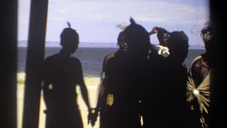 1969 Sudan frame showing silhouetted figures in the foreground and men in traditional costume near a sandy water edge in the background.
