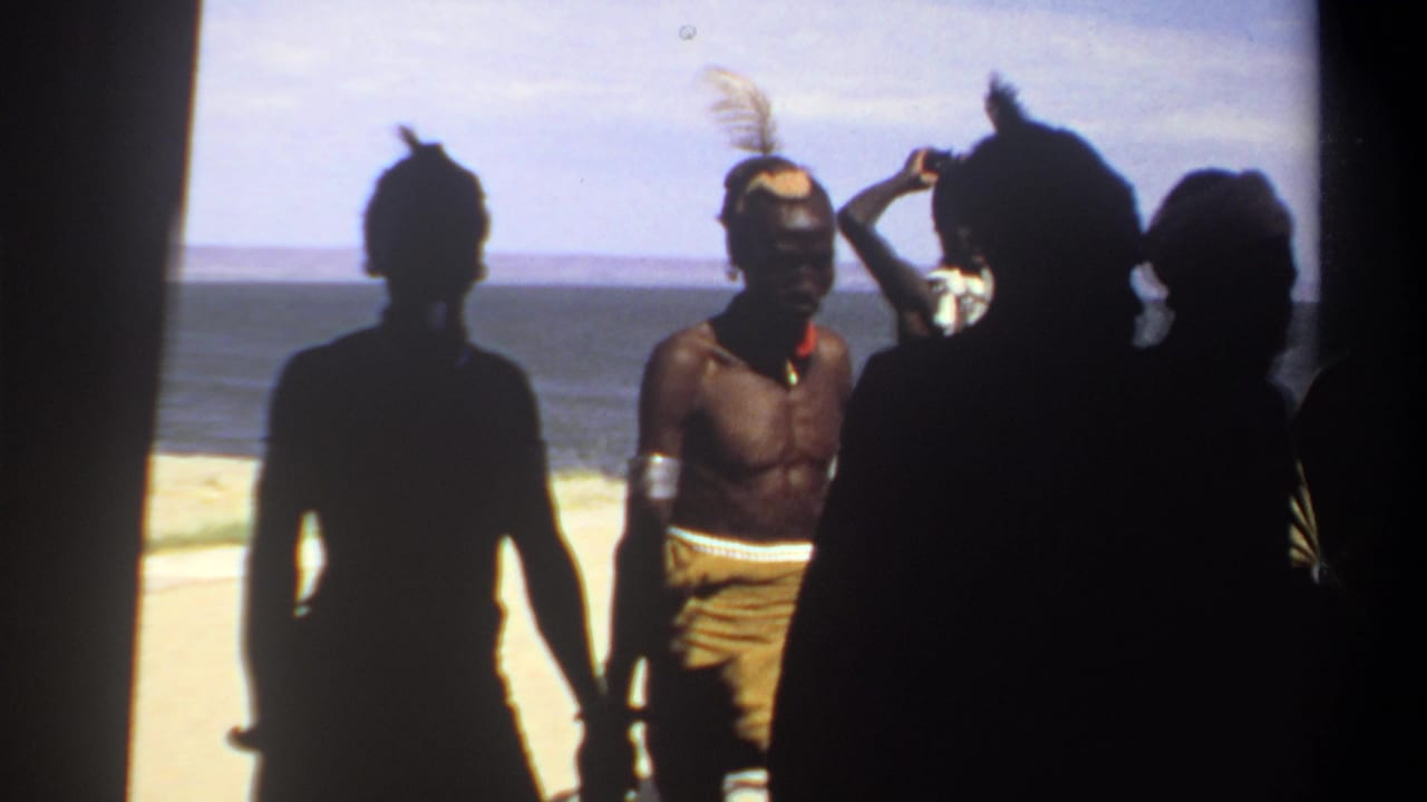 1969 Sudan home-movie frame showing men in feathered headdresses and bead necklaces on a bright sandy shoreline, with several figures in silhouette.