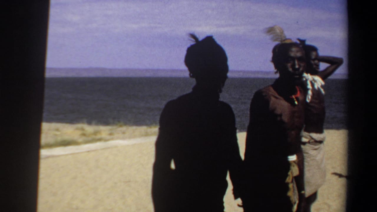 Two silhouetted figures stand near a man in a feathered headdress on the beach.