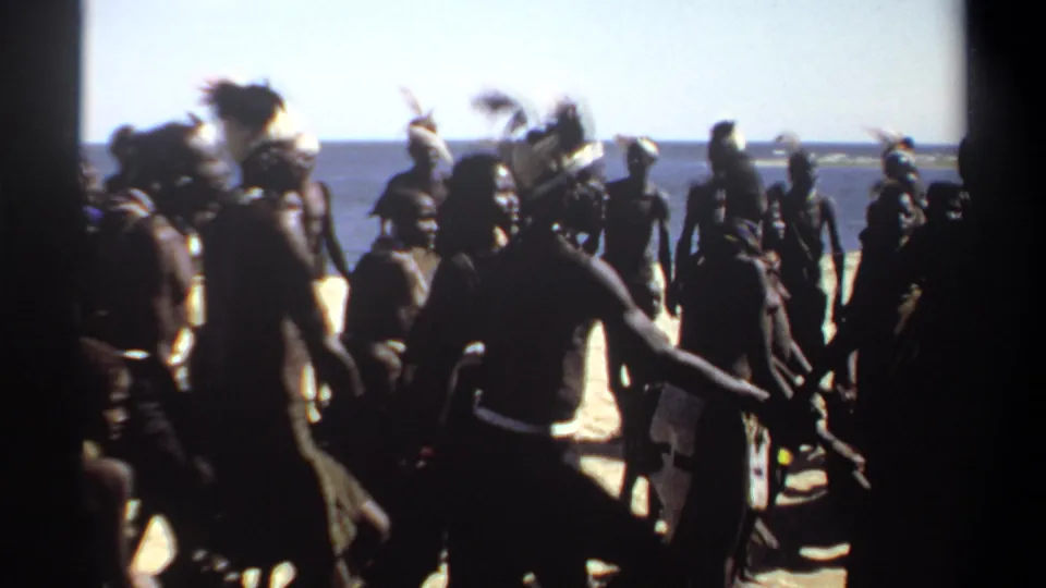 1969 Sudan home-movie frame showing a larger group of men in traditional attire and feathered headpieces gathered on a bright beach beside open water.