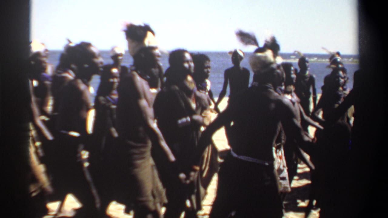 1969 Sudan 8mm frame showing silhouetted dancers on a sunlit beach, some wearing feathered headdresses, with the ocean behind them.