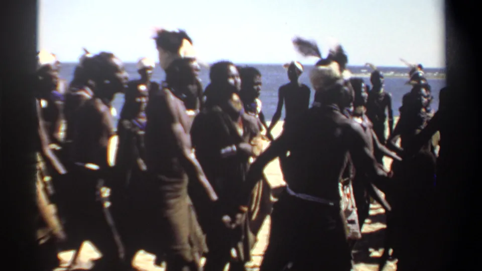 1969 Sudan 8mm frame showing silhouetted dancers on a sunlit beach, some wearing feathered headdresses, with the ocean behind them.