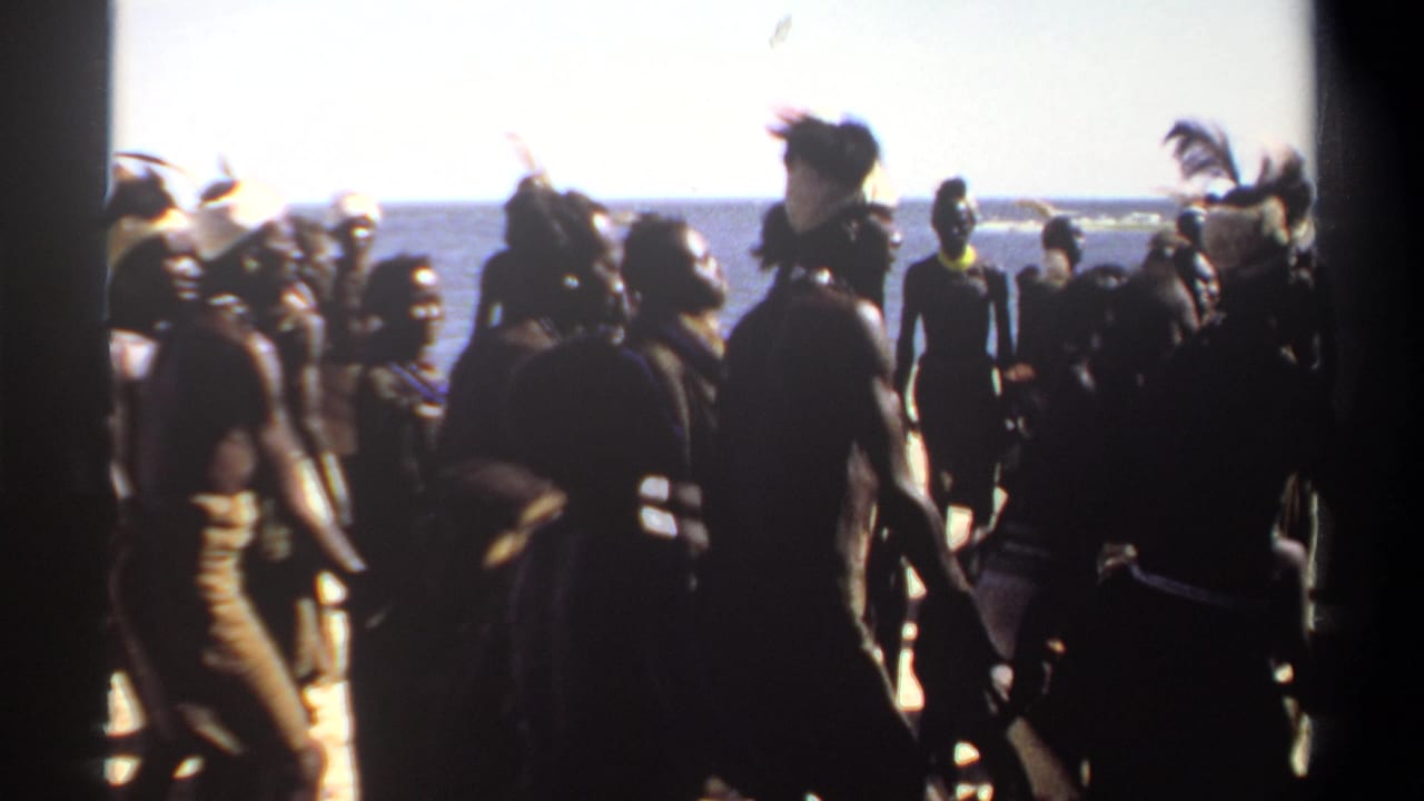 Participants in ceremonial dress move along a beach near the water.