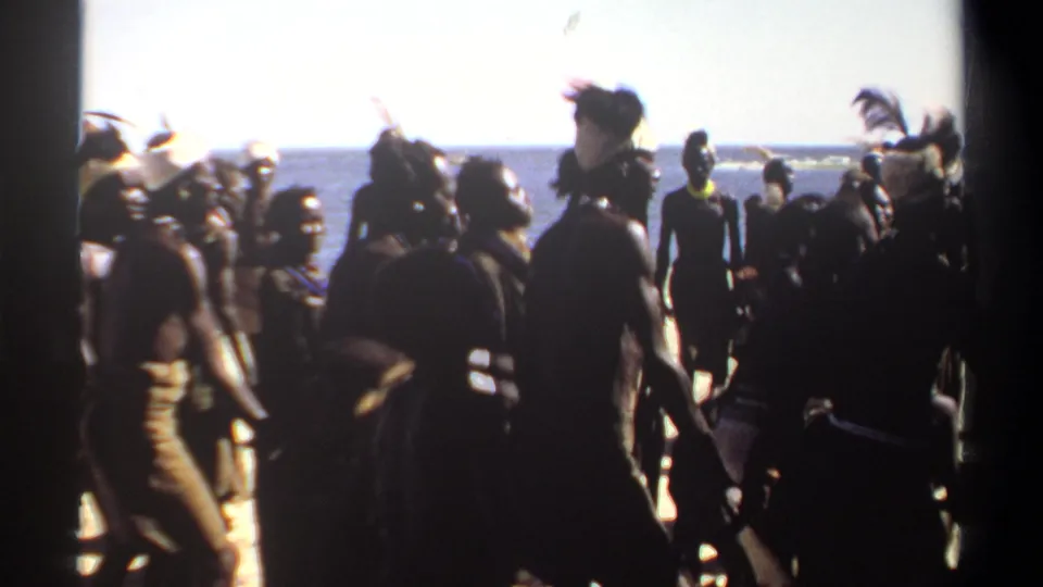 1969 Sudan 8mm frame with a beach procession or dance group, including feathered headwear and silhouetted figures against the sea.
