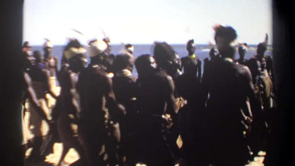 1969 Sudan 8mm frame looking through a crowd of dancers and onlookers toward the ocean during a shoreline ceremony.