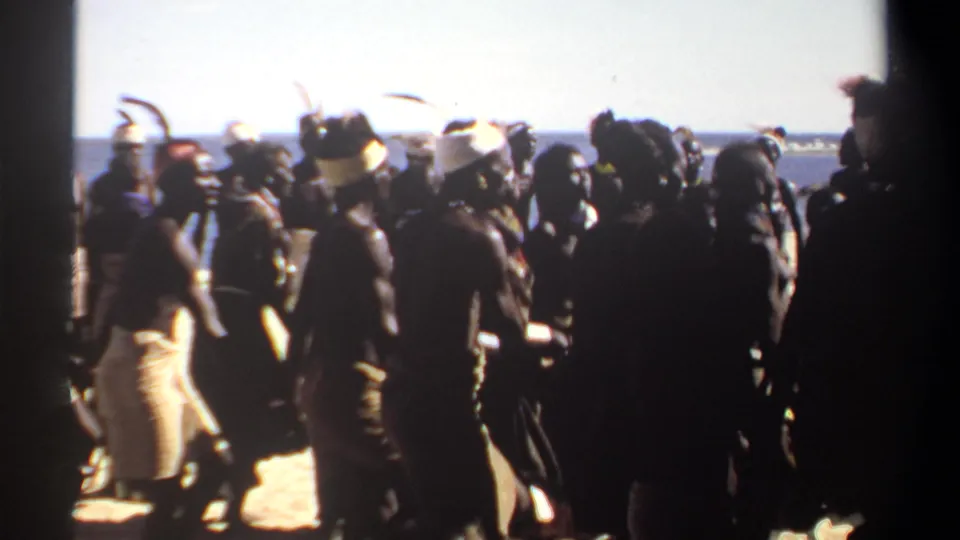 1969 Sudan 8mm frame showing silhouetted participants in a beach dance or ceremony, with feathered adornment and the sea in the distance.
