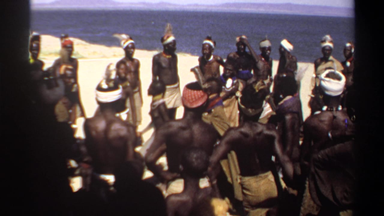 Men in a circle on sandy ground beside blue water, several wearing headwraps and wrapped cloths.