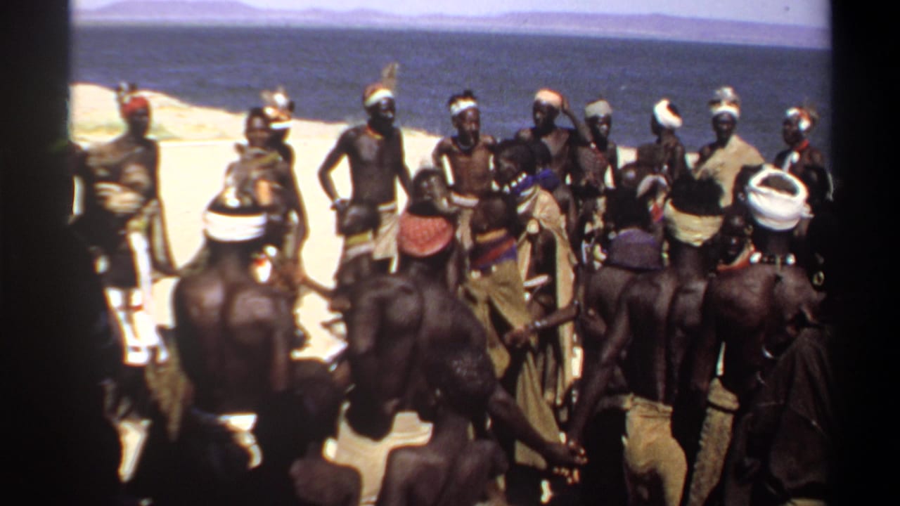 Group of men facing one another on sand, with water and a low horizon in the background.
