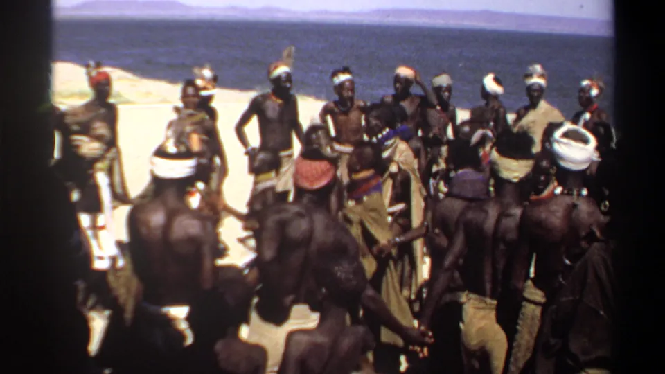 1969 Sudan home-movie frame of men in headwraps and cloth garments standing in a loose circle near water.