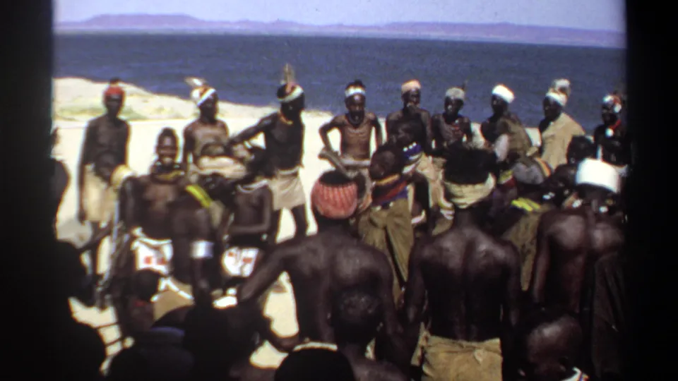1969 Sudan home-movie frame of men in a shoreline dance circle, with water and distant land visible beyond.