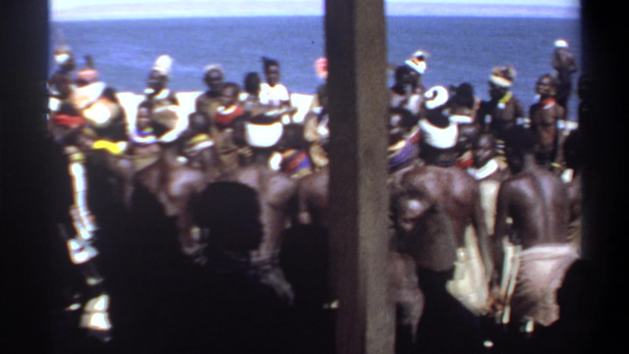 1969 Sudan home-movie frame showing a beachside crowd in ceremonial attire, partially obscured by wooden posts.