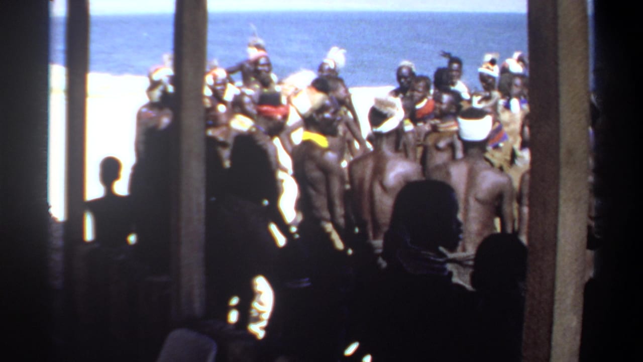 1969 Sudan frame of adults gathered on sand near blue water, with headdresses and wraps visible through shaded posts.