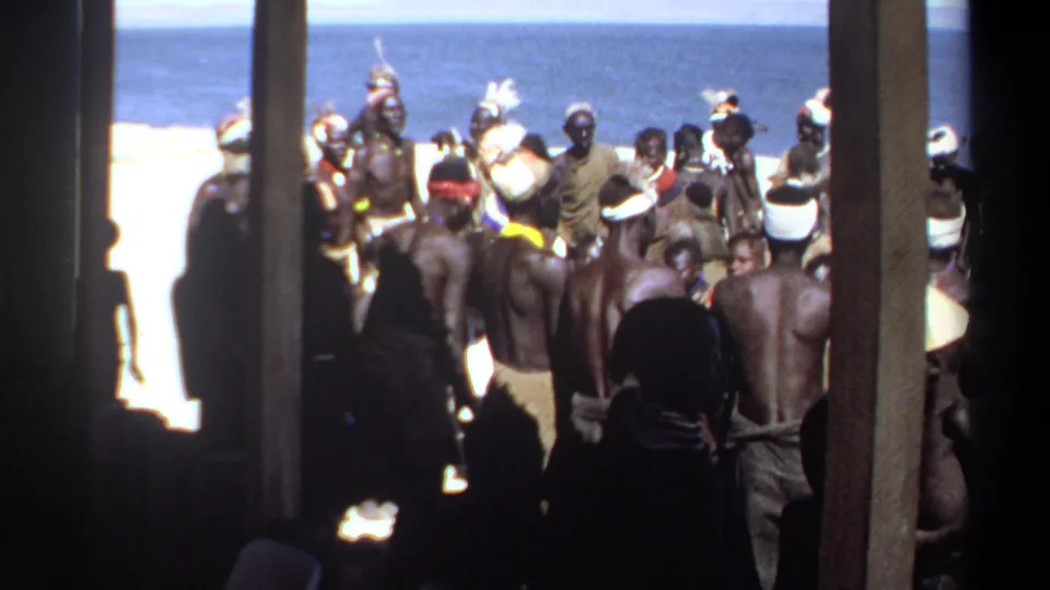 1969 Sudan home-movie view of a crowded shoreline gathering, filmed from a shaded position behind posts and onlookers.