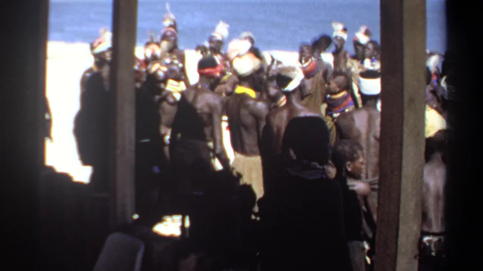 1969 Sudan frame showing a beachside cultural gathering with adults in headdresses and wraps, partly hidden by structural posts.