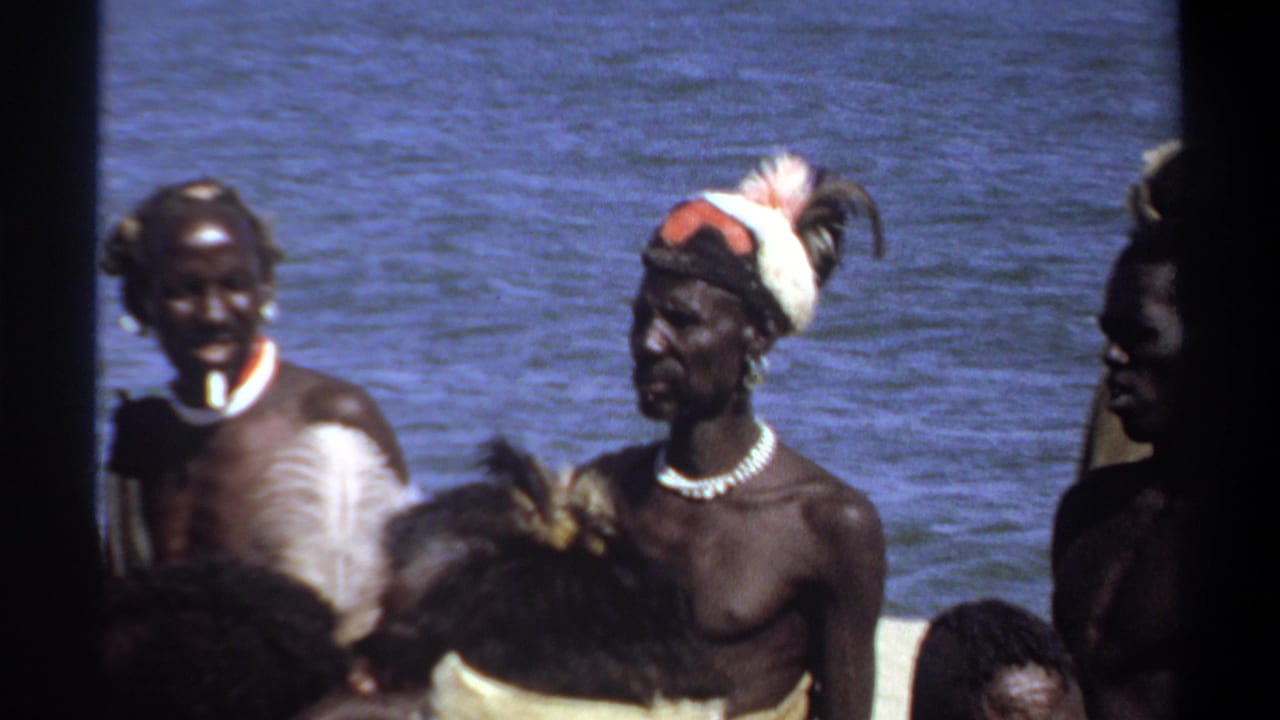 1969 Khartoum 8mm frame showing adult men in feathered headdresses, bead necklaces, and armbands near water.
