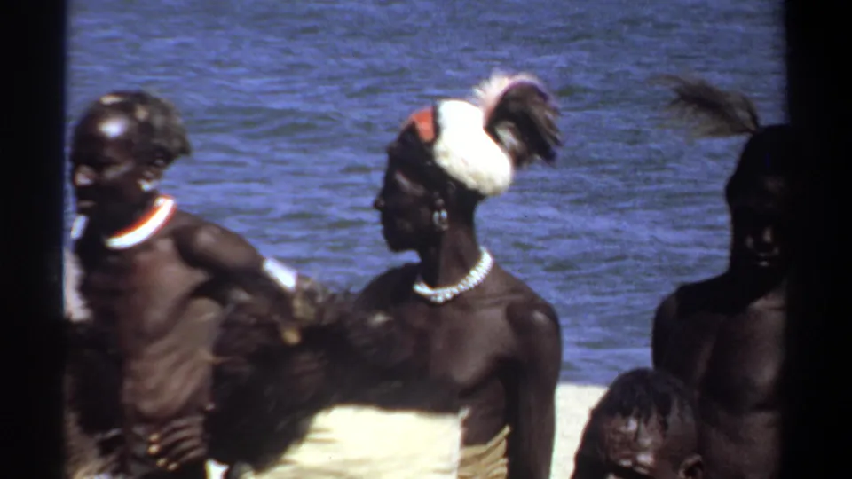 1969 Khartoum 8mm frame showing men in feathered headdresses and bead necklaces near water, with foreground crowding.