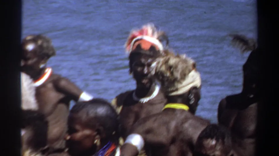 1969 Khartoum frame with a man in a feathered headdress facing the camera amid a riverside group.