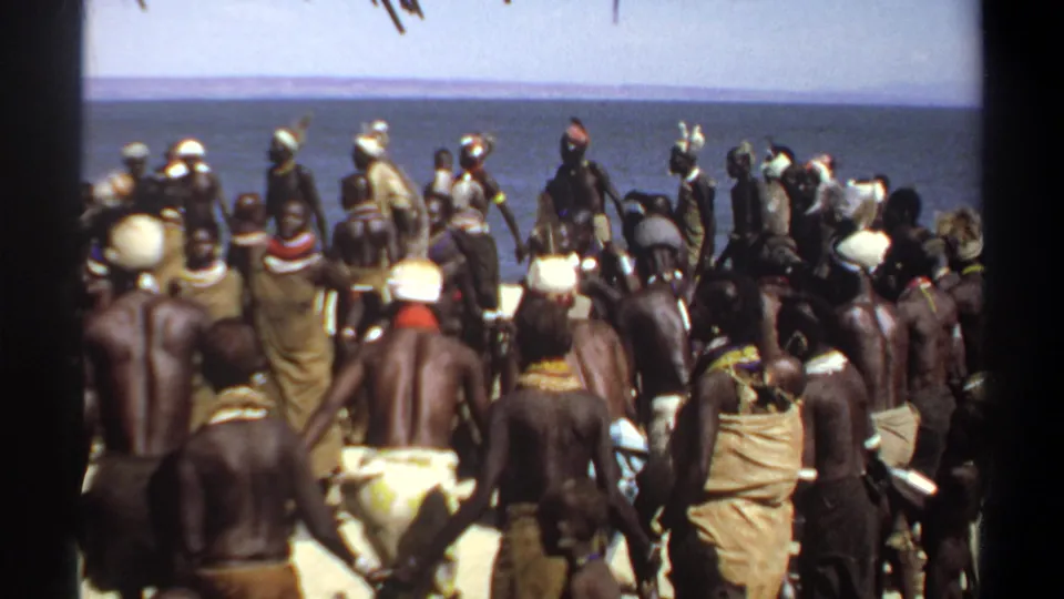1969 Sudan home-movie view of a large group gathered on a sandy beach, with water and a distant horizon behind them.
