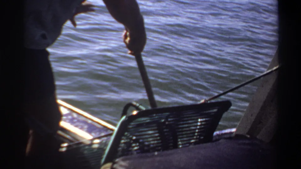 1969 Sudan home-movie frame of a man holding a fishing pole or net handle from a small boat on calm water.