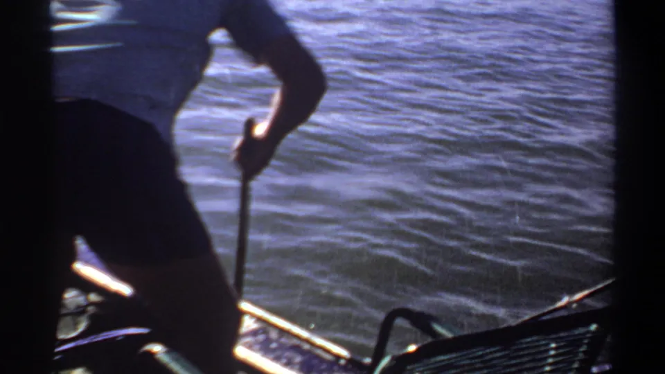 Backlit 1969 Sudan frame showing a man in shorts handling fishing gear from a small boat on rippled water.