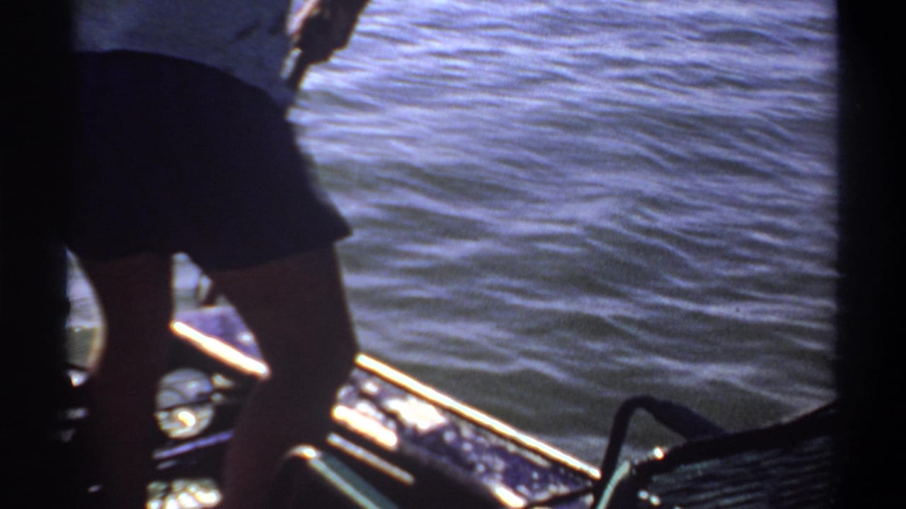 1969 Khartoum home-movie frame showing partial figures on a small boat near blue water, with sun glare, boat rails, and 8mm frame edges.