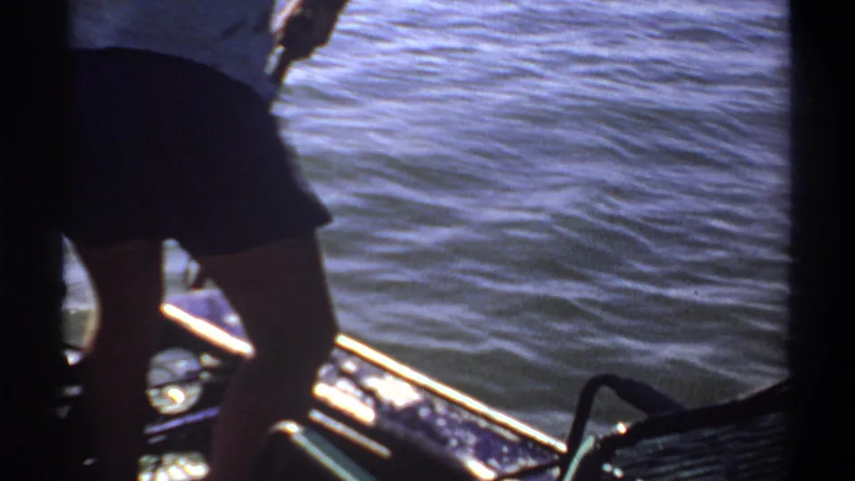 1969 Khartoum home-movie frame showing partial figures on a small boat near blue water, with sun glare, boat rails, and 8mm frame edges.