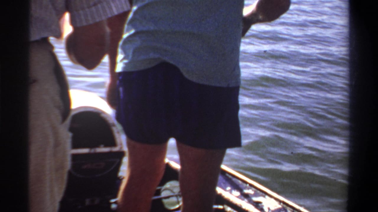 Standing person in a light shirt and dark shorts at the stern of a small boat on blue water.