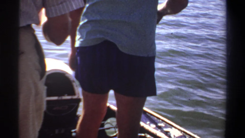 1969 Khartoum home-movie frame with a standing person in a light shirt and dark shorts near the stern of a small boat on bright blue water.