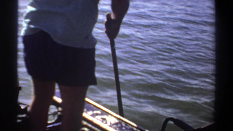 1969 Khartoum home-movie frame showing a person holding a paddle from a small boat, seen close to rippling blue water and boat rails.