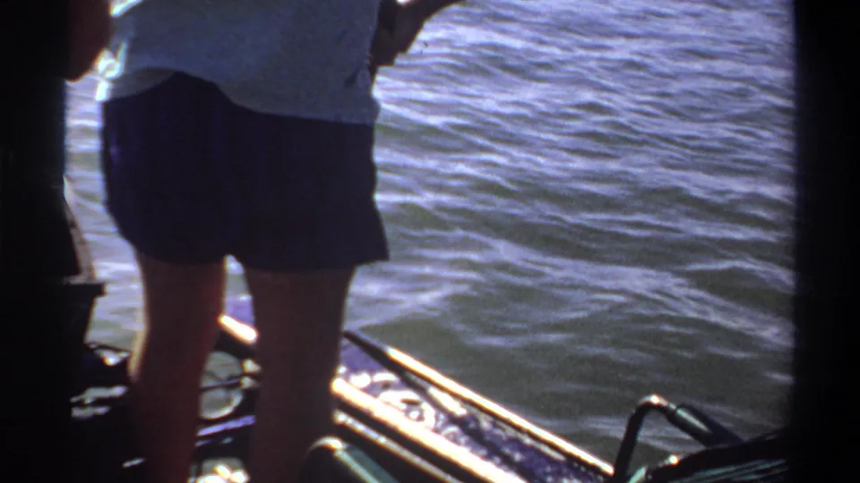 1969 Khartoum home-movie frame with a standing person on a small boat, rail details, and blue rippling water under bright sunlight.