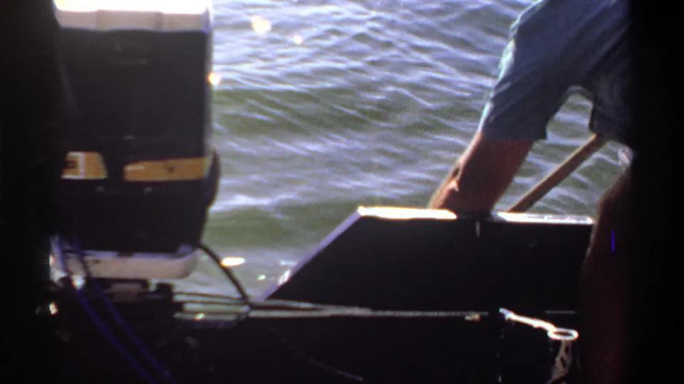 1969 Khartoum home-movie frame showing a person in a light blue shirt leaning by the stern as water passes behind the boat.
