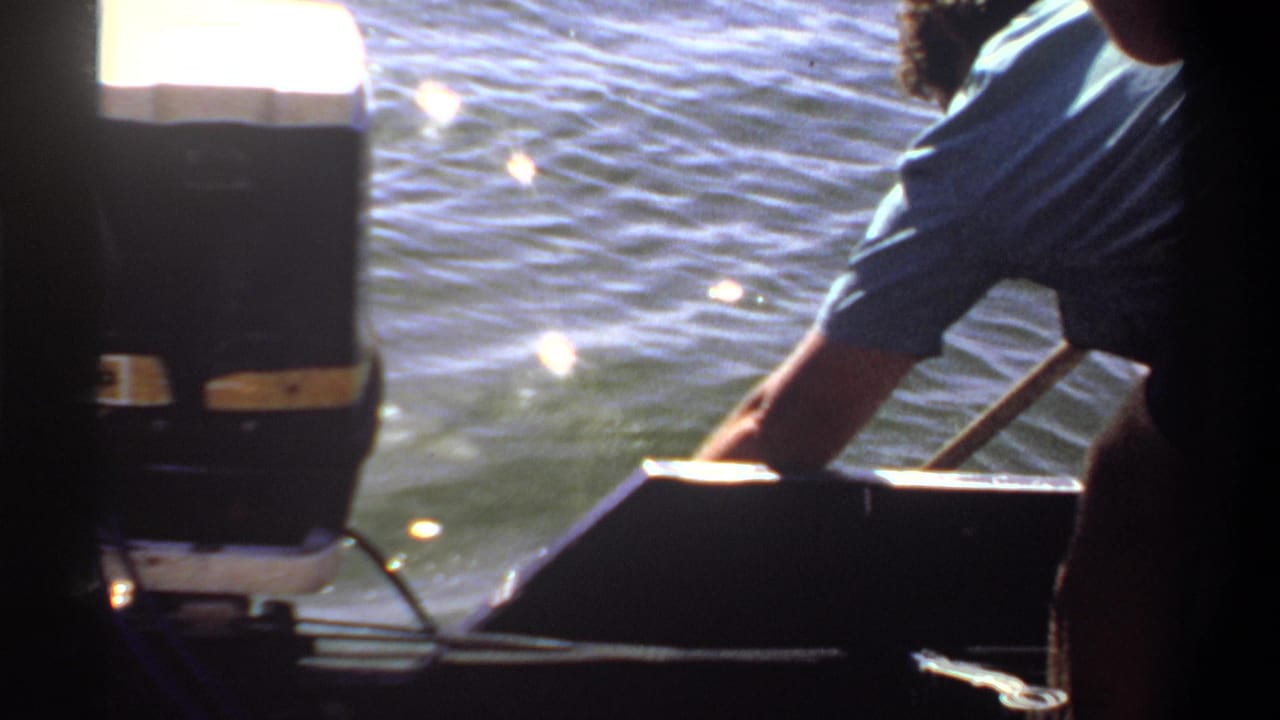 1969 Khartoum home-movie frame showing a man leaning over a small boat near an outboard motor, with sunlit water behind him.