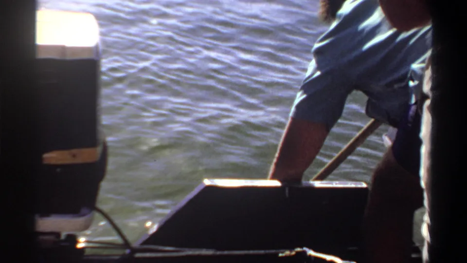 Home-movie frame from Khartoum in 1969 showing a man bent over a small boat with a long tool or pole near the water.