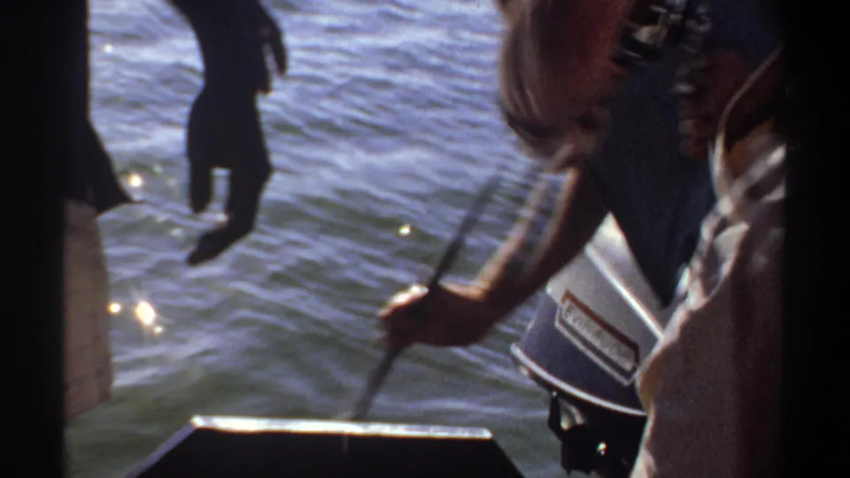1969 Khartoum fishing moment with a person leaning over the boat edge near an outboard motor as others stand nearby over the water.