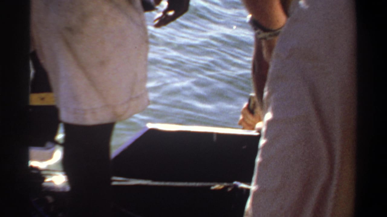 1969 Khartoum home-movie frame showing men in light clothing standing near the edge of a small boat with water behind them.