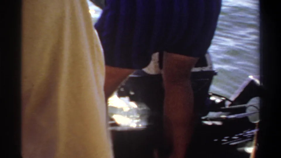 Close 1969 Khartoum boat view showing men's legs, dark shorts, bright water glare, and the boat edge during fishing activity.