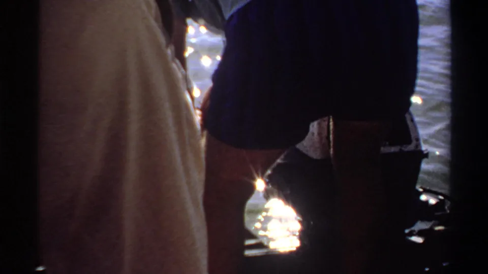 1969 Khartoum home-movie frame showing cropped figures in a boat, with shorts, light clothing, dark supports, and sunlit water reflections.
