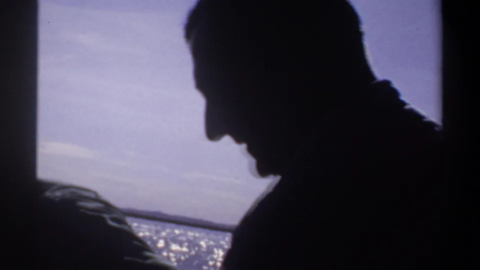 A silhouetted adult figure sits or leans on the boat, with shimmering water and pale sky behind them in the 1969 Sudan home movie.