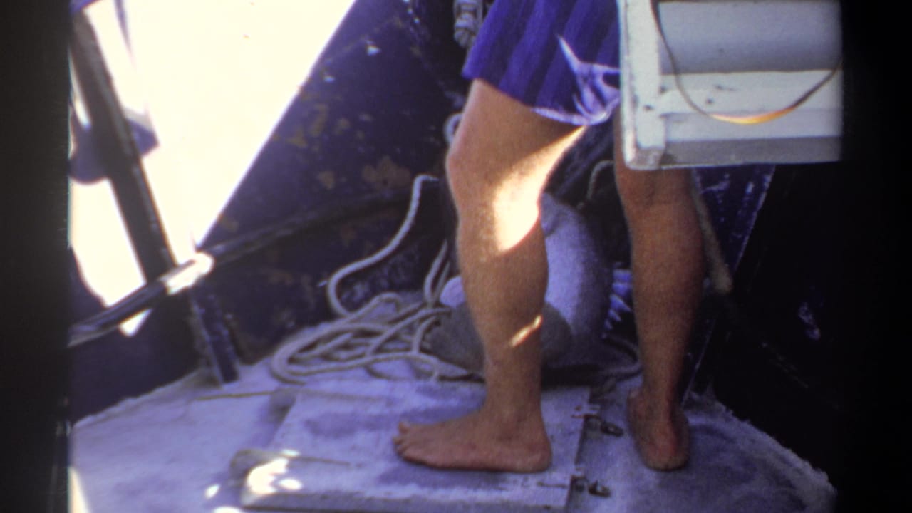 1969 Khartoum home-movie frame showing a barefoot fisherman in blue shorts standing on a boat deck beside coiled rope.